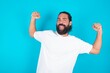 © Jihan - Yes I am winner. Portrait of charming delighted and excited young bearded man wearing white T-shirt over blue  raising up fist in triumph and victory smiling achieving success grinning from delight.