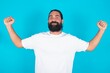 © Jihan - Waist up shot of young bearded man wearing white T-shirt over blue  raises arms to show muscles feels confident in victory, looks strong and independent, smiles positively at camera. Sport concept.