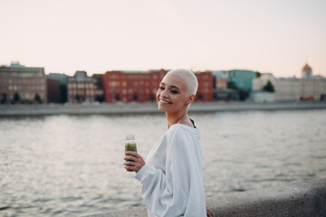 Wall Mural - Portrait of young smiling millenial european short haired woman with green smoothie. Beautiful happy blonde girl outdoor.