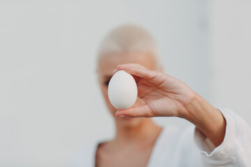 Wall Mural - Portrait of young smiling millenial european short haired woman hold egg in hand. Beautiful happy blonde girl outdoor.