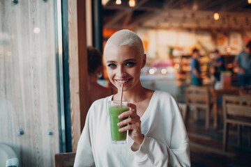 Wall Mural - Portrait of young smiling millenial european short haired woman with green smoothie at cafe. Beautiful happy blonde girl indoor.