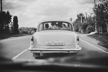 Happy Bride Kiss Groom, Newlywed Wedding Couple Is Driving A Retro Car On A Country Road For Honeymoon After The Ceremony. Way. The Best Day And Marriage. Just Married. Black And White Photo.