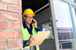 © Syda Productions - construction business and building concept - happy smiling male builder in helmet and safety west with clipboard calling on smartphone at window
