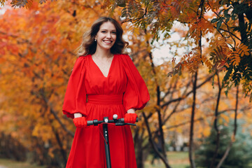 Wall Mural - Young woman in red dress riding electric scooter in autumn city. Alternative commuter transportation.