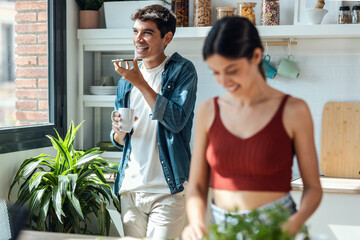 Couple in the kitchen preparing breakfast and sending voice message with mobile phone in the kitchen at home