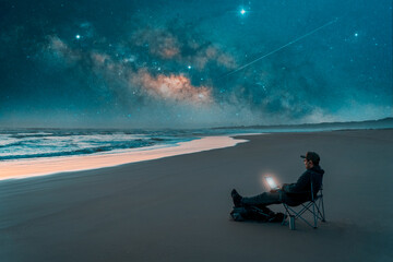 mature latin male sitting at night on the shore of the beach with a laptop and stars in the background