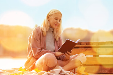 Mature woman reading interesting book near river on autumn day