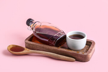 wooden board with bottle of maple syrup, bowl and spoon on pink background