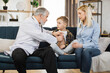 © sofiko14 - Preschool sick cute boy sitting with his mother on the couch while caring experienced nice mature male doctor listening his heartbeat during home visit, front view.