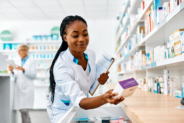 Wall Mural - Young smiling black pharmacist going through inventory in pharmacy.