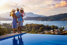 A Family On Summer Holidays Stands By The Swimming Pool And Enjoys The Beautiful Sunset Behind The Mountains And The Sea