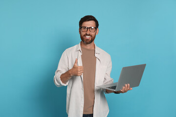 Handsome man with laptop showing thumb up gesture on light blue background