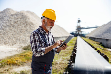 Heavy industry and factories. A senior factory worker with a helmet on his head stands next to a conveyor belt and analyzes production on his tablet.