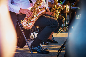 A group of people from a musical brass band who sit on chairs and play music, jazz on golden brass pipes, sit outdoors in the rotunda. Shiny patent leather shoes. The baluster is blurred