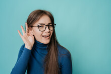 I Can’t Hear You! Pretty Caucasian Young Female In Glasses Holding Hand Above Ear Asking To Be Louder Over Turquoise Backdrop. Pretty Student Girl Wants To Go To Party, Mockup For Breaking News.