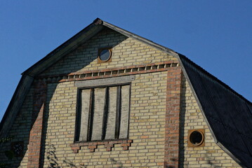 Sticker - one attic of an old brown abandoned brick private house with a window boarded up with plywood under a gray slate roof against a blue sky