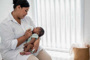 Asian mother feeding milk from bottle milk to her 1-month-old baby black skin newborn son, is Half-Nigerian Half-Thai, to family and food for infant concept.