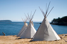 Three Teepees Are Lined Up In A Row In Front Of Water At A Summer Camp.
