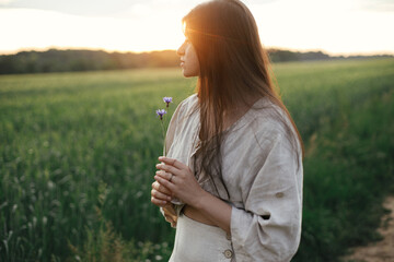 Wall Mural - Woman relaxing in wheat field in warm sunset light. Stylish young female in rustic dress holding wildflowers in hands in evening summer countryside. Tranquil atmospheric moment