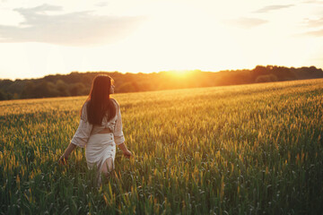 Wall Mural - Woman walking in wheat field in warm sunset light. Stylish young female in rustic dress holding wildflowers in hands and relaxing in evening summer countryside. Tranquil atmospheric moment