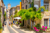 Fototapeta Uliczki - A picturesque main street through the historic medieval town of Saint-Remy de Provence, France, with the colorful shops and cafes and the clock tower in view on a summer day.