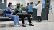 © DC Studio - African american patient and female nurse talking about healthcare in waiting room lobby, doing consultation in hospital reception. Diverse people doing checkup examination at facility. Tripod shot.