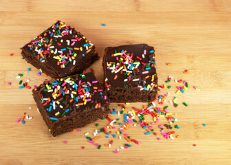 Freshly baked brownies with chocolate frosting and multicolored sprinkles are displayed on a wooden cutting board.