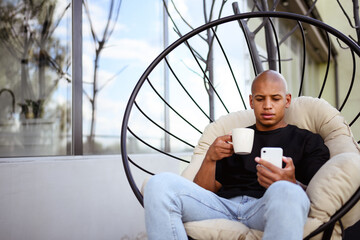 Young african american man holding coffee and using smartphone on armchair on balcony 