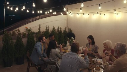 Wall Mural - Family and friends celebrating at dinner on a rooftop terrace. Multiethnic group of people dining on the balcony on a special evening to celebrate friendship and family love and relationships