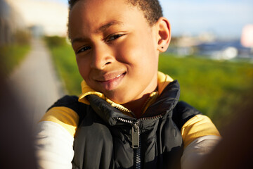 Wall Mural - Closeup cropped self-portrait of cute black kid boy taking selfie walking down big city, spending leisure time playing with front camera on his new smartphone. Children and technology