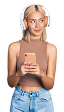 Beautiful young blonde woman using smartphone wearing headphones smiling looking to the side and staring away thinking.