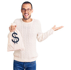 Young handsome man holding money bag with dollar symbol celebrating victory with happy smile and winner expression with raised hands