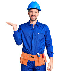 Canvas Print - Young handsome man wearing worker uniform and hardhat smiling cheerful presenting and pointing with palm of hand looking at the camera.