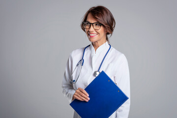 Smiling female doctor standing at isolated grey background and holding clipboard in her hand