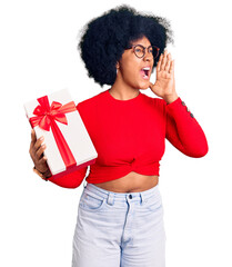 Wall Mural - Young african american girl holding gift shouting and screaming loud to side with hand on mouth. communication concept.