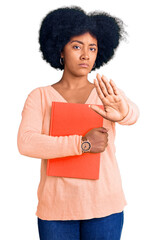 Wall Mural - Young african american girl holding book with open hand doing stop sign with serious and confident expression, defense gesture