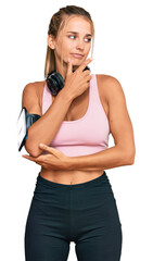 Wall Mural - Young blonde woman wearing gym clothes and using headphones with hand on chin thinking about question, pensive expression. smiling with thoughtful face. doubt concept.