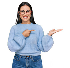 Young hispanic woman wearing casual clothes and glasses amazed and smiling to the camera while presenting with hand and pointing with finger.