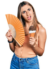 Poster - Hispanic young woman holding hand fan eating ice cream in shock face, looking skeptical and sarcastic, surprised with open mouth