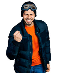 Wall Mural - Young hispanic man with beard wearing snow wear and sky glasses angry and mad raising fist frustrated and furious while shouting with anger. rage and aggressive concept.