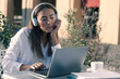 © Paulista - Focused young woman in headphones attending a webinar through laptop sitting in an outdoor cafe.
