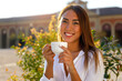 © Paulista - Smiling young woman in white shirt looking at camera sipping cappuccino or tea  in street cafe. Urban lifestyles concept
