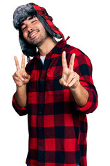 Sticker - Hispanic young man with beard wearing fluffy earmuff hat smiling looking to the camera showing fingers doing victory sign. number two.