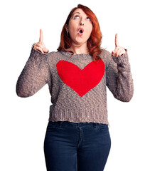 Wall Mural - Young beautiful redhead woman wearing casual heart sweater amazed and surprised looking up and pointing with fingers and raised arms.