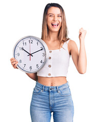 Canvas Print - Young beautiful girl holding big clock screaming proud, celebrating victory and success very excited with raised arms