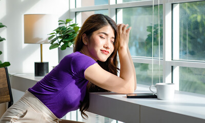 Asian millennial young beautiful female housewife taking break resting relaxing sitting on chair near glass windows smelling drinking hot coffee on table counter bar in living room at home on weekend