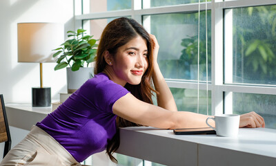 Asian millennial young beautiful female housewife taking break resting relaxing sitting on chair near glass windows smelling drinking hot coffee on table counter bar in living room at home on weekend