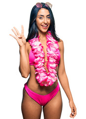 Wall Mural - Beautiful hispanic woman wearing bikini and hawaiian lei showing and pointing up with fingers number four while smiling confident and happy.