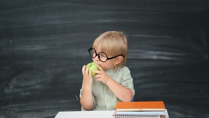 Back to school. Happy smiling pupil in round glasses sitting at the desk. Child eating green apple in the classroom with blackboard on background. Kid boy from primary school. First day of fall.