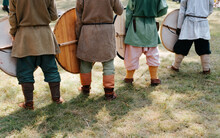 Back View Of Medieval Soldiers With Shields Standing In A Row On The Grass On The Battlefield. Cropped Image